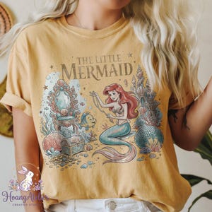 May include: Mustard yellow t-shirt featuring a graphic of Ariel from The Little Mermaid. The design includes Ariel, Flounder, a treasure chest, and the text "The Little Mermaid". The design is in shades of blue, pink, and gold.