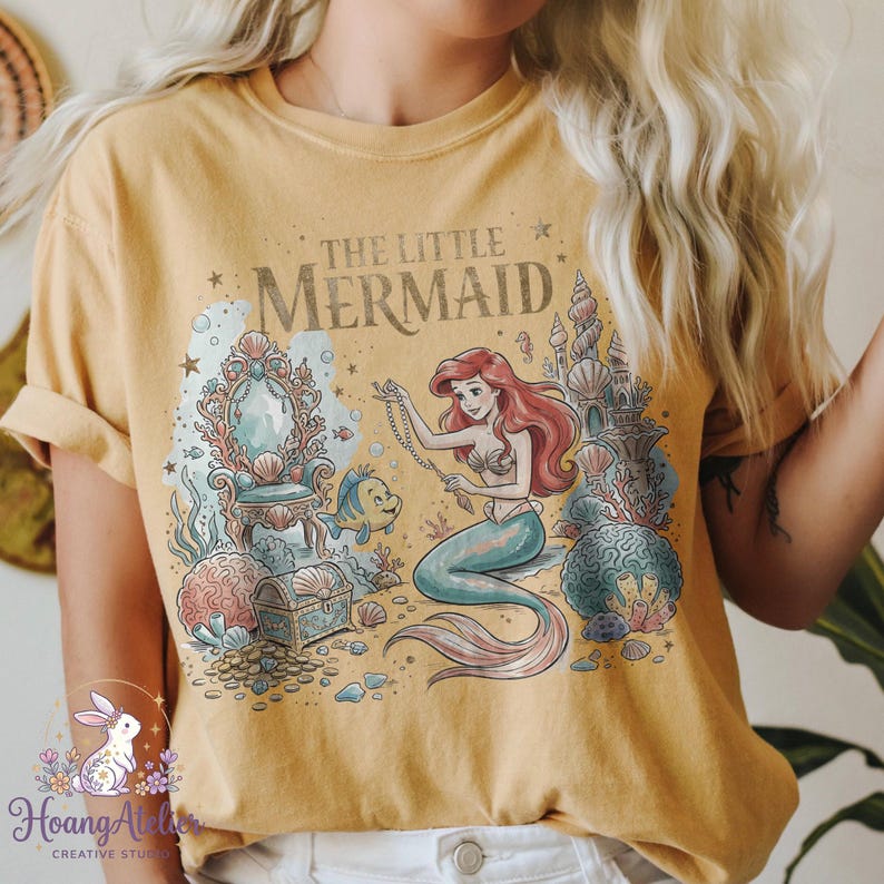 May include: Mustard yellow t-shirt featuring a graphic of Ariel from The Little Mermaid. The design includes Ariel, Flounder, a treasure chest, and the text "The Little Mermaid". The design is in shades of blue, pink, and gold.