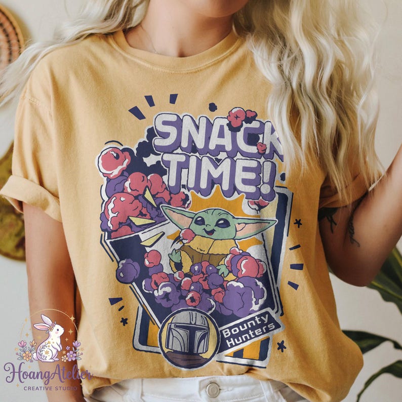 Puede incluir: Camiseta de color amarillo mostaza con un dise&ntilde;o de Grogu, tambi&eacute;n conocido como Baby Yoda, con el texto "SNACK TIME!" y "Bounty Hunters". El dise&ntilde;o incluye palomitas de ma&iacute;z y un casco mandaloriano.
