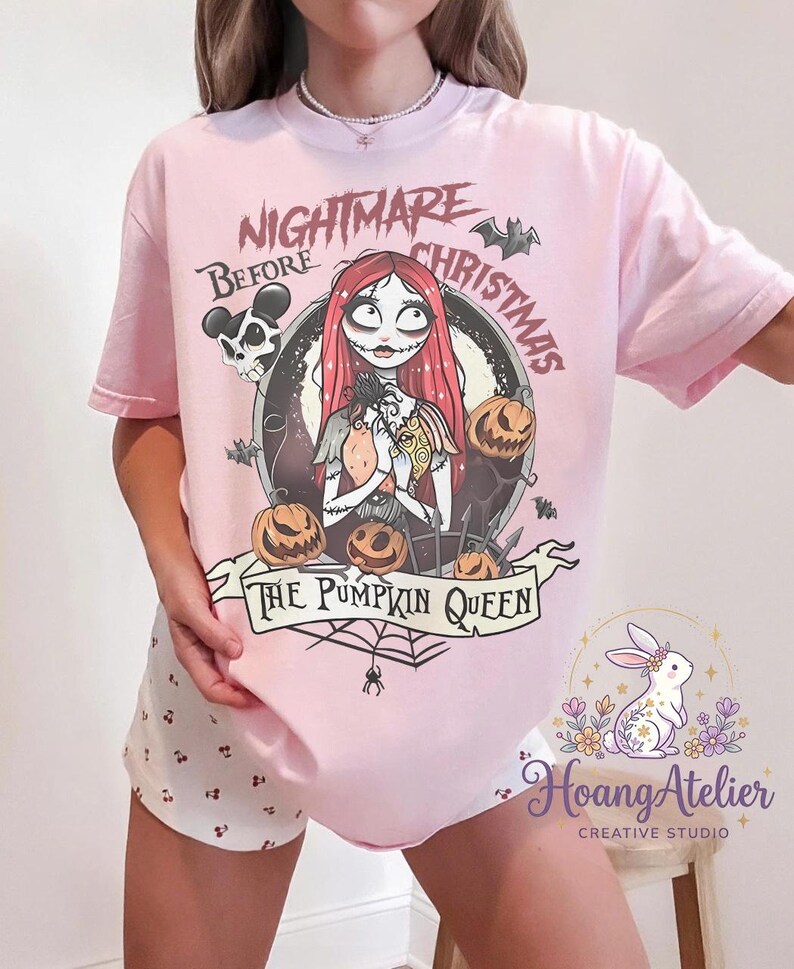 May include: A light pink t-shirt featuring a graphic from "The Nightmare Before Christmas." The design includes Sally, pumpkins, bats, and the text "Nightmare Before Christmas" and "The Pumpkin Queen."