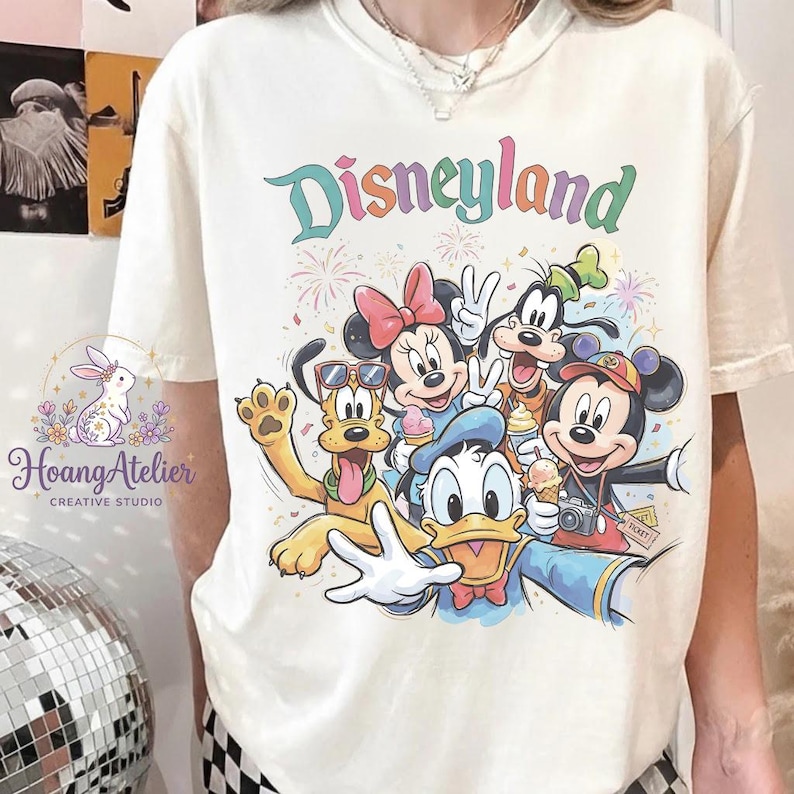 May include: Cream-colored t-shirt featuring the word "Disneyland" in colorful lettering above a graphic of Disney characters including Mickey Mouse, Minnie Mouse, Donald Duck, Goofy, and Pluto. The design includes fireworks and ice cream cones.