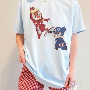 May include: Light blue t-shirt featuring cartoon characters dressed as superheroes. One character is in a red Iron Man suit, and the other is in a Captain America costume. The shirt is paired with red and white checkered shorts.