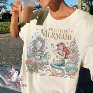 May include: Cream-colored t-shirt featuring a colorful illustration of Ariel from The Little Mermaid. The design includes a mermaid, a treasure chest, and the text "THE LITTLE MERMAID". The shirt has short sleeves and a crew neck.