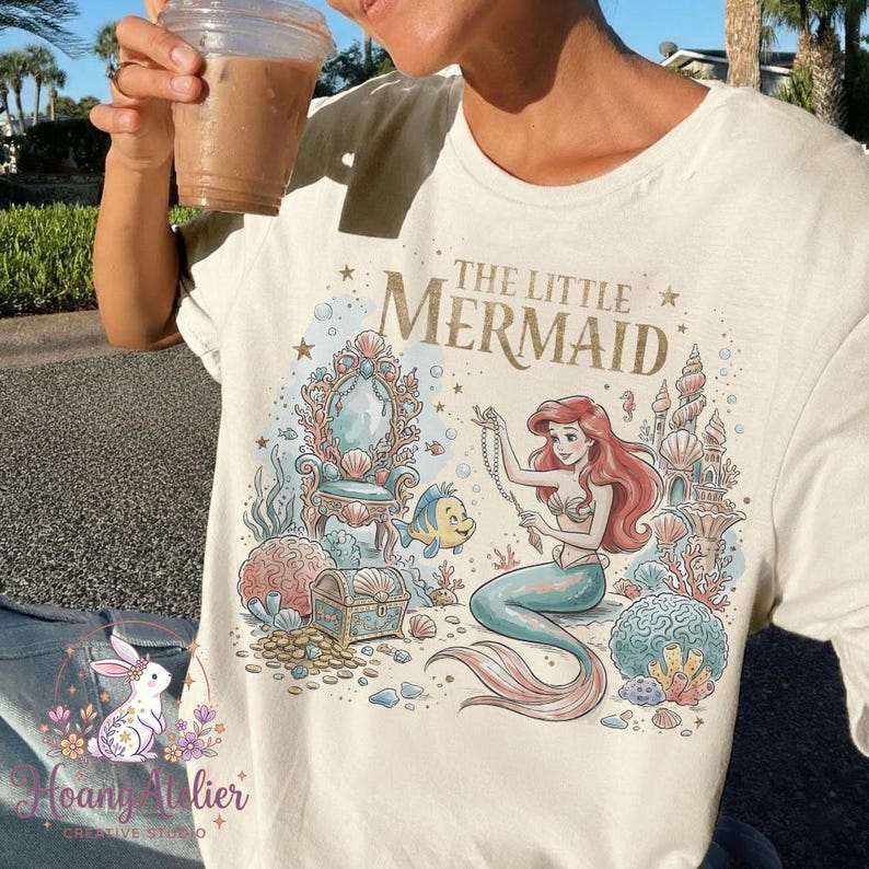 May include: Cream-colored t-shirt featuring a colorful illustration of Ariel from The Little Mermaid. The design includes a mermaid, a treasure chest, and the text "THE LITTLE MERMAID". The shirt has short sleeves and a crew neck.