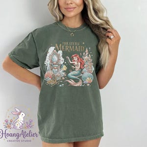 May include: Olive green t-shirt featuring a colorful graphic of Ariel from "The Little Mermaid." The design includes a mermaid, castle, and treasure chest. The shirt also has the text "The Little Mermaid."