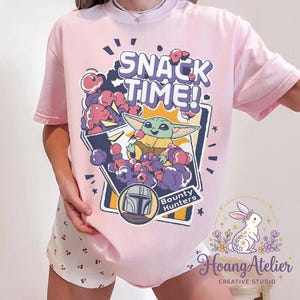 Puede incluir: Camiseta rosa claro con un gr&aacute;fico de Grogu de The Mandalorian y el texto "SNACK TIME!". El dise&ntilde;o incluye un casco de cazarrecompensas y las palabras "Bounty Hunters".