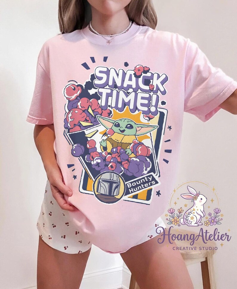Puede incluir: Camiseta rosa claro con un gr&aacute;fico de Grogu de The Mandalorian y el texto "SNACK TIME!". El dise&ntilde;o incluye un casco de cazarrecompensas y las palabras "Bounty Hunters".