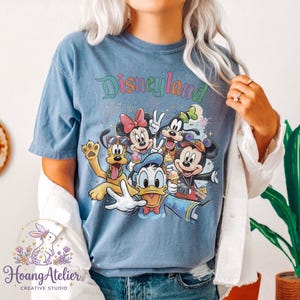May include: Blue t-shirt featuring the word "Disneyland" in colorful script above a graphic of Mickey Mouse, Minnie Mouse, Donald Duck, Goofy, and Pluto. The design has a vintage, distressed look.