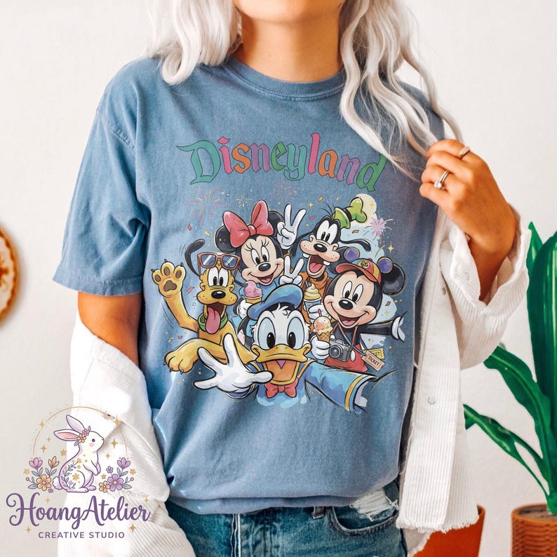 May include: Blue t-shirt featuring the word "Disneyland" in colorful script above a graphic of Mickey Mouse, Minnie Mouse, Donald Duck, Goofy, and Pluto. The design has a vintage, distressed look.
