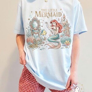 May include: Light blue t-shirt featuring a colorful illustration of Ariel from The Little Mermaid. The graphic includes the text "THE LITTLE MERMAID" in gold lettering, a mermaid, Flounder, and underwater elements. The shirt is worn with red and white checkered shorts.
