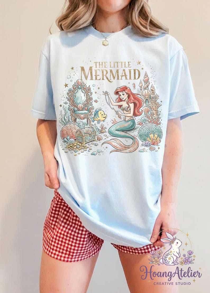 May include: Light blue t-shirt featuring a colorful illustration of Ariel from The Little Mermaid. The graphic includes the text "THE LITTLE MERMAID" in gold lettering, a mermaid, Flounder, and underwater elements. The shirt is worn with red and white checkered shorts.