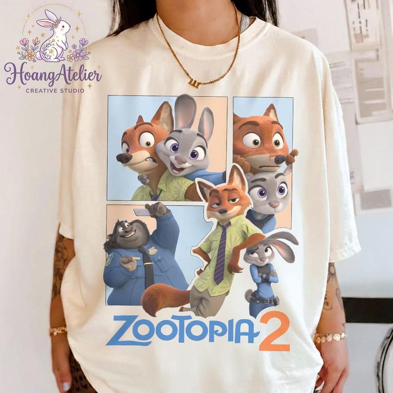 K&ouml;nnte beinhalten: Cremefarbenes T-Shirt mit einer farbenfrohen Grafik von Charakteren aus dem Animationsfilm "Zootopia 2". Das Design enth&auml;lt Bilder eines Fuchses, eines Kaninchens und anderer Charaktere im Comic-Stil. Die Worte "Zootopia 2" sind blau gedruckt.