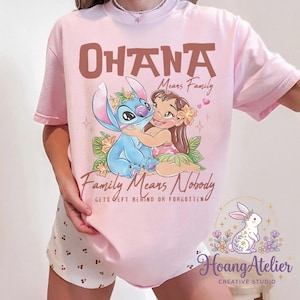 Op de afbeelding: Lichtroze T-shirt met een afbeelding van Stitch en Lilo uit de film Lilo & Stitch. De tekst "OHANA Means Family" en "Family Means Nobody Gets Left Behind or Forgotten" staat boven en onder de afbeelding. Het shirt heeft ook het Hoang Atelier logo.