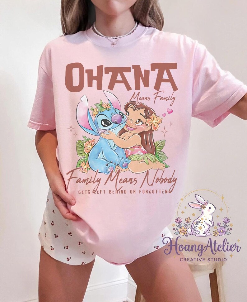 Op de afbeelding: Lichtroze T-shirt met een afbeelding van Stitch en Lilo uit de film Lilo & Stitch. De tekst "OHANA Means Family" en "Family Means Nobody Gets Left Behind or Forgotten" staat boven en onder de afbeelding. Het shirt heeft ook het Hoang Atelier logo.