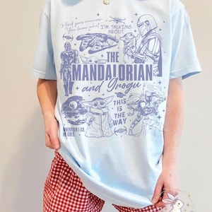 Puede incluir: Camiseta azul claro con ilustraciones de "The Mandalorian", incluyendo al Mandaloriano y Grogu, y el texto "The Mandalorian and Grogu". La camiseta tambi&eacute;n tiene las frases "Mind your manners" y "This is the way".