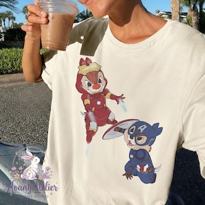 May include: Cream-colored t-shirt featuring cartoon characters dressed as superheroes. One character is in a red Iron Man suit, and the other is in a blue Captain America suit. The shirt has a Hoang Atelier logo.