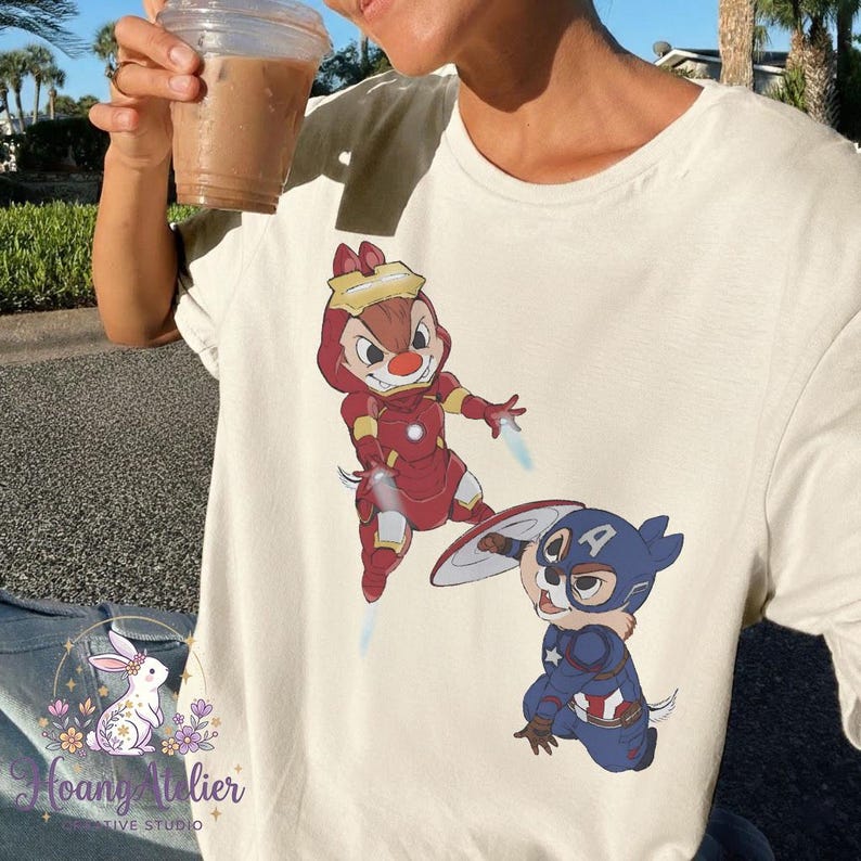 May include: Cream-colored t-shirt featuring cartoon characters dressed as superheroes. One character is in a red Iron Man suit, and the other is in a blue Captain America suit. The shirt has a Hoang Atelier logo.