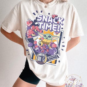 Puede incluir: Camiseta color crema con un gr&aacute;fico de Grogu de The Mandalorian y el texto "SNACK TIME!". El dise&ntilde;o incluye bocadillos de dibujos animados morados y rosas y las palabras "Bounty Hunters".