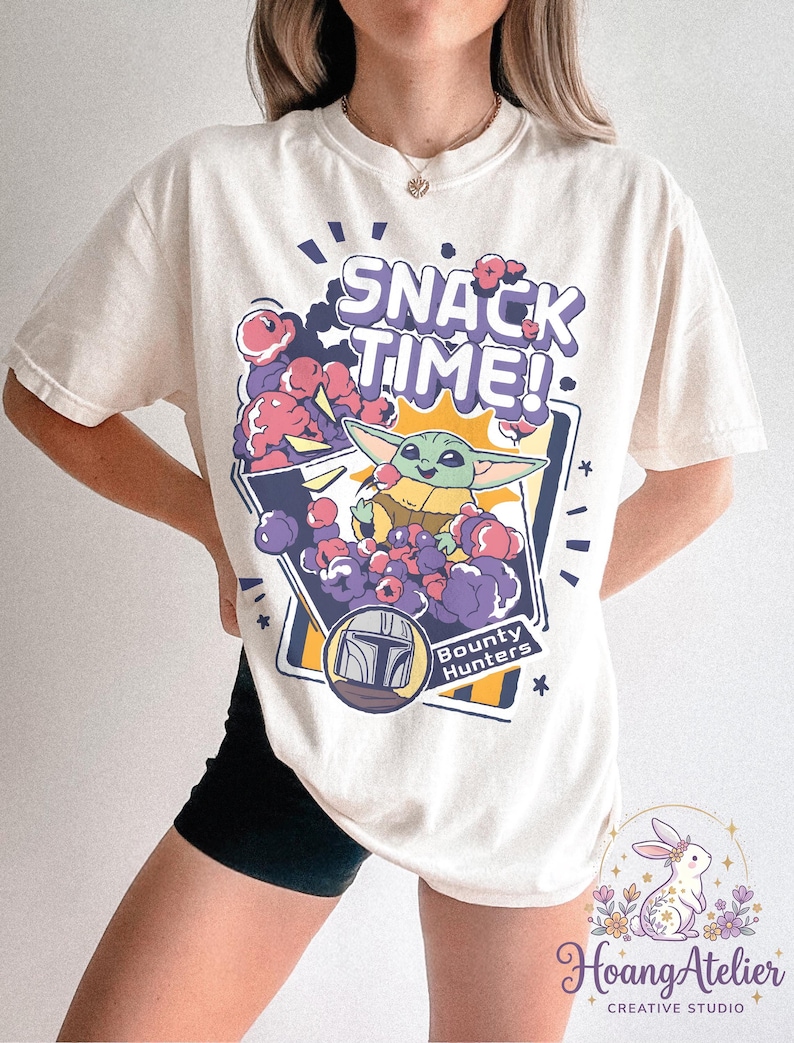 Puede incluir: Camiseta color crema con un gr&aacute;fico de Grogu de The Mandalorian y el texto "SNACK TIME!". El dise&ntilde;o incluye bocadillos de dibujos animados morados y rosas y las palabras "Bounty Hunters".