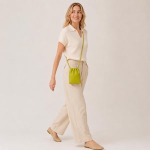 Può includere: Una polo a maniche corte color crema a coste, pantaloni ampi color crema e una borsa a tracolla color verde lime con chiusura a coulisse. La borsa presenta una tracolla lunga e sottile in tinta, completata da scarpe basse color beige.