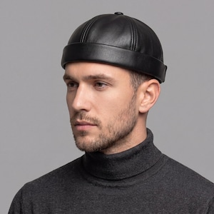 Puede incluir: Gorra de cuero negro con corona redondeada y ala corta. La gorra tiene costuras visibles y un pequeño botón en la parte superior. La persona lleva un jersey de cuello alto gris oscuro.