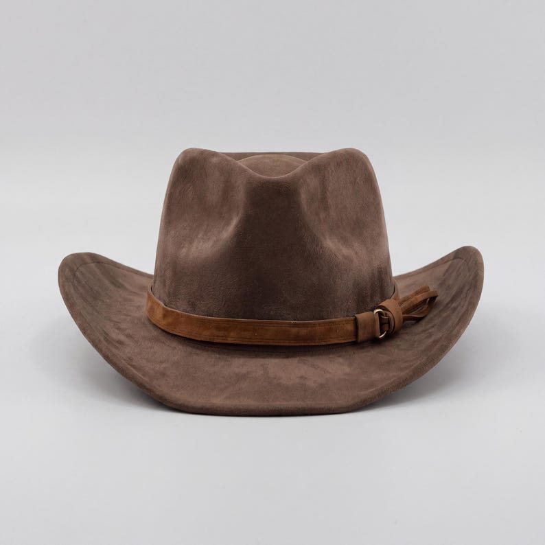 May include: A brown suede cowboy hat with a wide brim and a pinched crown. The hat features a brown leather band around the base of the crown and a decorative leather tassel. The hat is a neutral color and is suitable for various occasions.