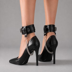 May include: A pair of black high-heeled pumps featuring a pointed toe and a slim stiletto heel. Each shoe includes a black ankle strap with a silver-toned buckle and a silver-toned metal chain that connects the ankle strap to the side of the shoe.