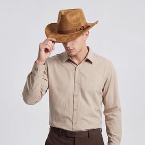 May include: A brown suede cowboy hat with a leather band and buckle detail. The hat has a wide brim and a classic western style. The person in the image is wearing a tan button-down shirt.