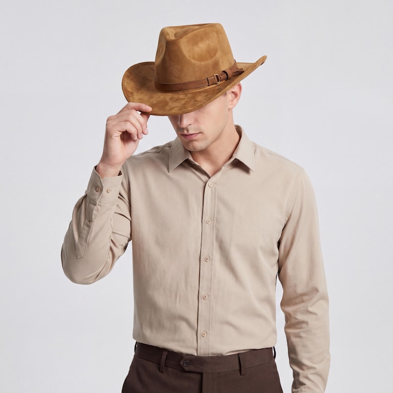 May include: A brown suede cowboy hat with a leather band and buckle detail. The hat has a wide brim and a classic western style. The person in the image is wearing a tan button-down shirt.