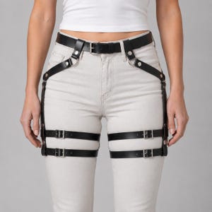 May include: A black leather-like leg harness featuring a waist belt connected to two thigh straps with silver-toned metal buckles, rivets, and O-rings. The harness is worn over light-colored denim jeans and a plain white cropped top.