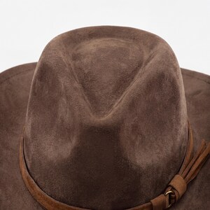 May include: A close-up of a brown fedora hat with a suede-like texture. The hat features a brown leather band around the crown and a decorative tassel. The hat is set against a white background.