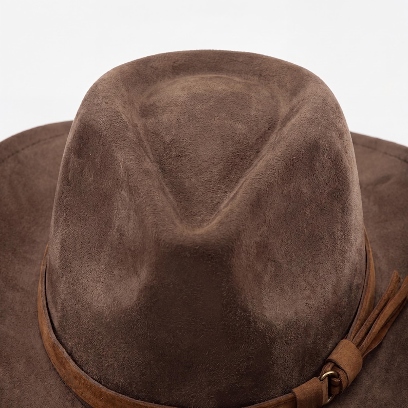 May include: A close-up of a brown fedora hat with a suede-like texture. The hat features a brown leather band around the crown and a decorative tassel. The hat is set against a white background.