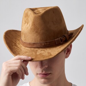 May include: A brown suede cowboy hat with a leather band and buckle detail. The hat has a classic brim and a slightly indented crown. The hat is a warm, earthy tone, perfect for adding a touch of Western style to any outfit.
