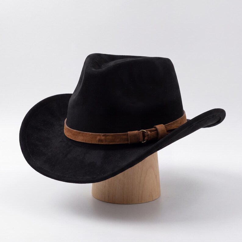 May include: A black cowboy hat with a brown leather band and buckle. The hat is displayed on a wooden hat stand. The hat has a wide brim and a classic western style.