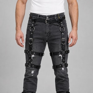 May include: Distressed charcoal gray denim jeans featuring a black leather harness system. The harness includes adjustable straps, silver-toned metal buckles, and circular rings that wrap around the thighs and connect to a waist belt.