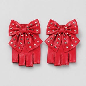 May include: A pair of bright red fingerless gloves featuring a large, prominent bow on the back of each hand. The bows are decorated with numerous small, round silver-toned metal studs arranged in a pattern across the surface of the red material.