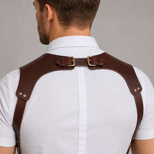 Könnte beinhalten: Ein dunkelbraunes Schulterholster aus Leder über einem weißen kurzärmeligen Hemd. Das Holster besteht aus zwei breiten Schulterriemen, die in der Mitte des Rückens durch einen horizontalen Ledersteg mit zwei Messingschnallen verbunden sind.