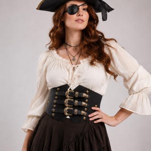 May include: A black corset-style belt featuring three horizontal straps with ornate gold-toned buckles and studs. The belt is worn over a cream-coloured long-sleeved blouse with a ruffled neckline and a dark brown skirt, paired with a black pirate hat.