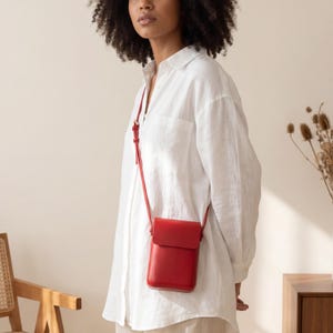 Op de afbeelding: Een rode leren crossbody tas met een rechthoekige vorm en een klepsluiting. De tas heeft een lange, dunne riem en wordt over de schouder gedragen. De tas is ongeveer 15 cm hoog en 10 cm breed.