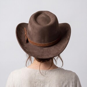May include: A brown suede cowboy hat with a brown leather band and a decorative leather tie. The hat has a wide brim and a rounded crown. The hat is worn by a person wearing a light-colored shirt.