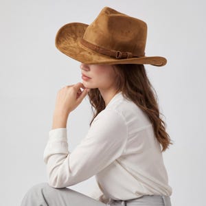 May include: A brown suede cowboy hat with a wide brim and a brown leather band. The hat has a classic western style and is suitable for outdoor activities. The hat is worn by a person wearing a white shirt.