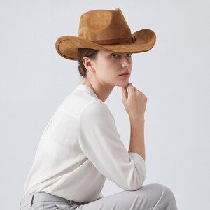 May include: A brown suede cowboy hat with a curved brim and a brown band. The hat is worn by a person wearing a white button-down shirt and gray pants. The hat's brim measures approximately 15 inches in diameter.
