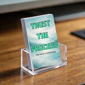 Peut inclure: Un jeu de cartes dans un support en acrylique transparent. Chaque carte présente un fond de ciel bleu et blanc avec les mots "TRUST THE PROCESS" en vert et noir. Le texte "The best is comming" est également visible.