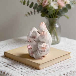 May include: A white and pink fox ornament with closed eyes, holding a pink gem, sits on a closed, aged book. The ornament has textured fur and delicate features. A vase of flowers is in the background.