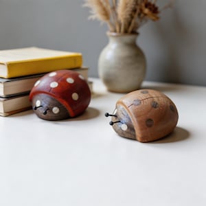 May include: Two wooden ladybug figurines. One is red with white spots, the other is brown with darker brown spots. Both have small black antennae and are set on a white surface. A stack of books and a vase with dried flowers are in the background.