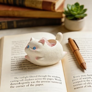May include: A white ceramic cat-shaped planter with pink accents on its ears and paws, and blue eyes. The planter is filled with pink stones. It is sitting on an open book, with a wooden pen nearby. A small succulent plant is in the background.