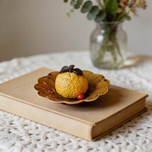 May include: A ceramic orange fruit sculpture with brown leaves sits in a decorative, scalloped-edge dish. The dish and fruit are on a closed book. A small red berry is next to the fruit. The fruit is yellow with a textured surface.