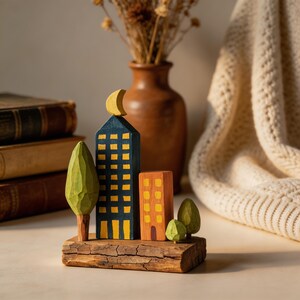 Peut inclure: Sculpture de paysage urbain en bois avec un grand bâtiment bleu et des fenêtres jaunes, un croissant de lune, un bâtiment orange et un arbre vert. Les bâtiments sont montés sur un morceau de bois, créant une scène urbaine miniature.