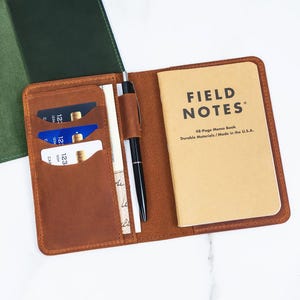 May include: A tan leather notebook cover featuring three card slots, a vertical pocket, a pen loop with a black pen, and a tan Field Notes memo book. The cover is open, revealing the interior organization. A separate dark green leather cover is visible.