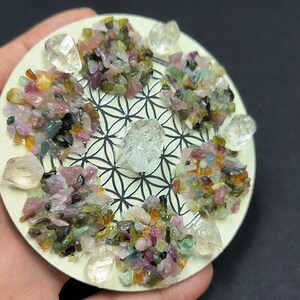 May include: A circular, cream-coloured decorative item featuring a black and white geometric design. It is embellished with clusters of colourful, crushed gemstones and clear quartz crystals. The gemstones include shades of pink, green, yellow, and blue.